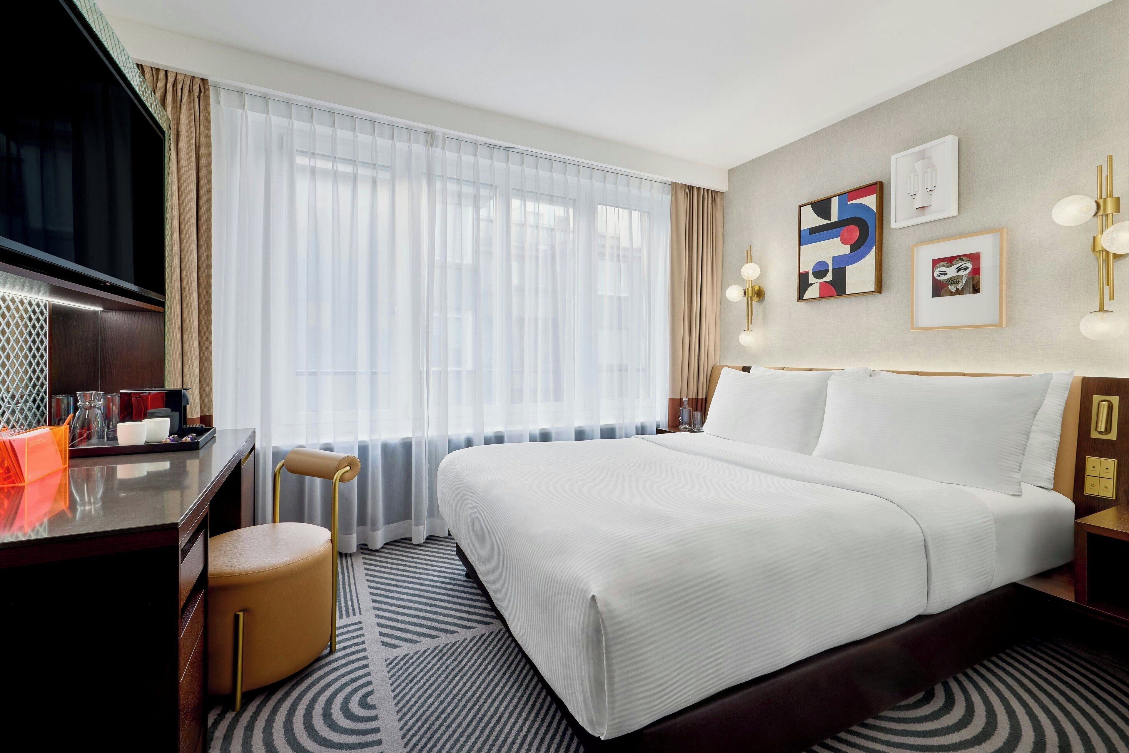 A hotel room with a large bed, a desk with a TV, a chair, and a window with sheer curtains.