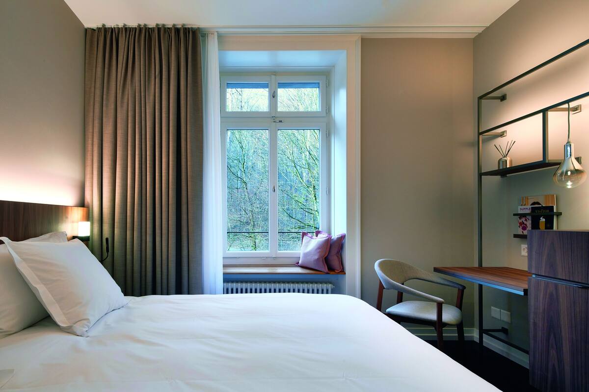 Une chambre d’hôtel avec un lit, une fenêtre avec des rideaux, un bureau, une chaise et un réfrigérateur.