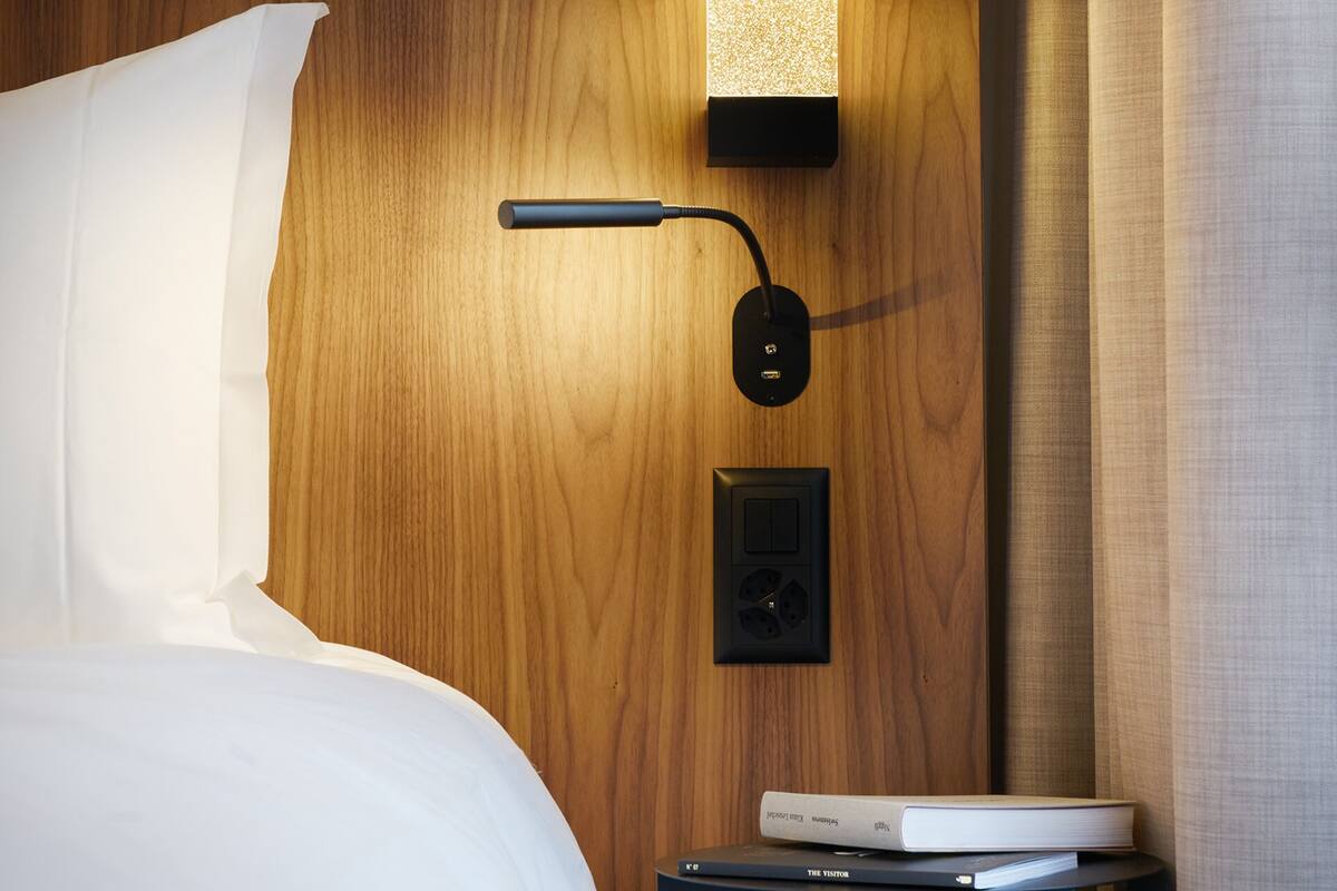 Un lit bien fait, avec du linge de lit blanc, une tête de lit en bois, une table de chevet avec une lampe et un petit appareil électronique posé sur la table.