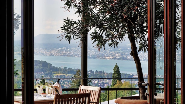 A balcony with a wooden chair, a table with wine glasses, and a view of a coastal town and mountains. - Sorell Boutique-Hotel Zürichberg (Zürich)