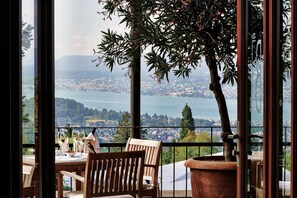 Een balkon met een houten stoel, een tafel met wijnglazen en uitzicht op een kustplaatsje en bergen.