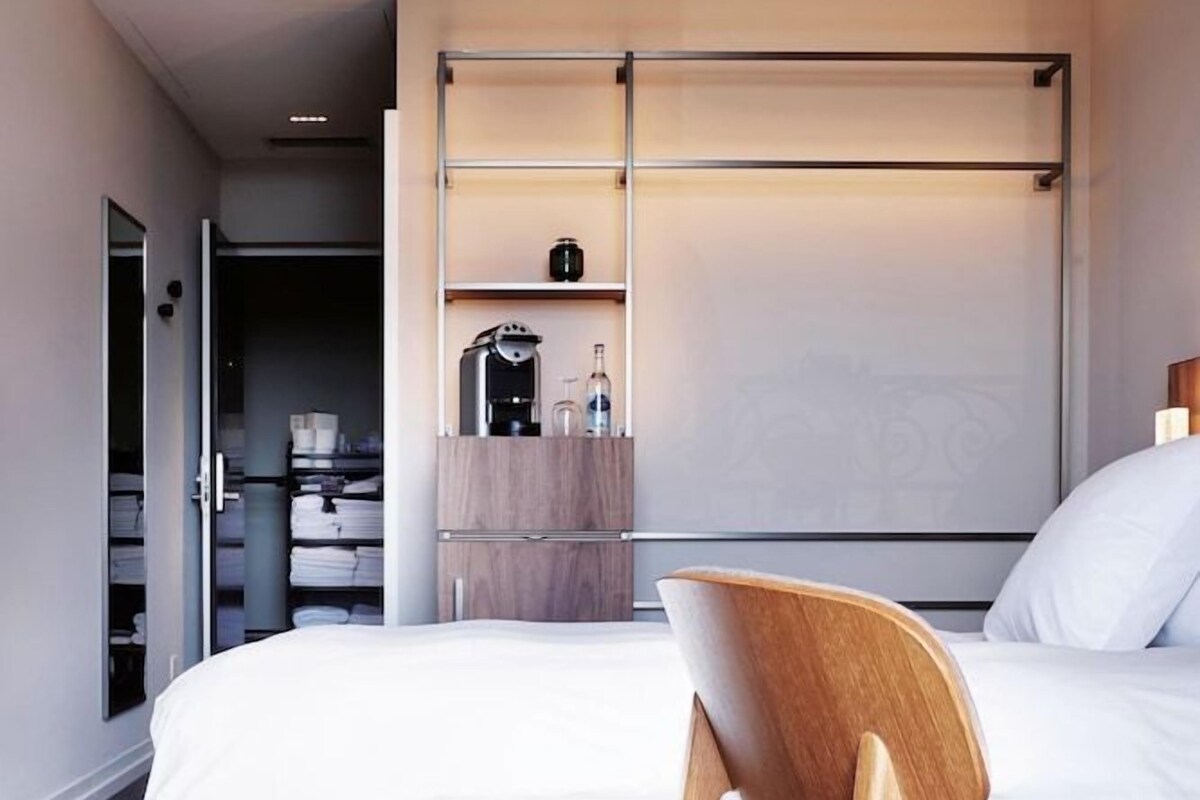 A modern hotel room with a bed, a wooden chair, a wardrobe, and a small kitchenette area.