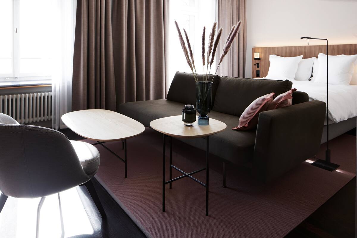 Une chambre d’hôtel moderne, dotée d’un canapé, de deux tables rondes, d’un vase agrémenté d’éléments décoratifs et d’un lit recouvert de linge de maison blanc.