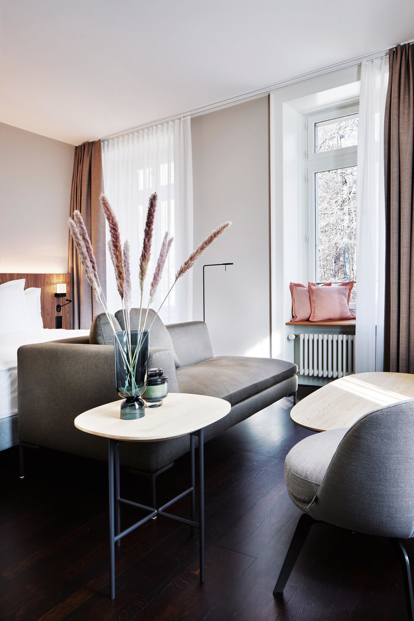 Une chambre d’hôtel moderne avec un canapé, une petite table, une chaise, une fenêtre avec des rideaux et un radiateur.