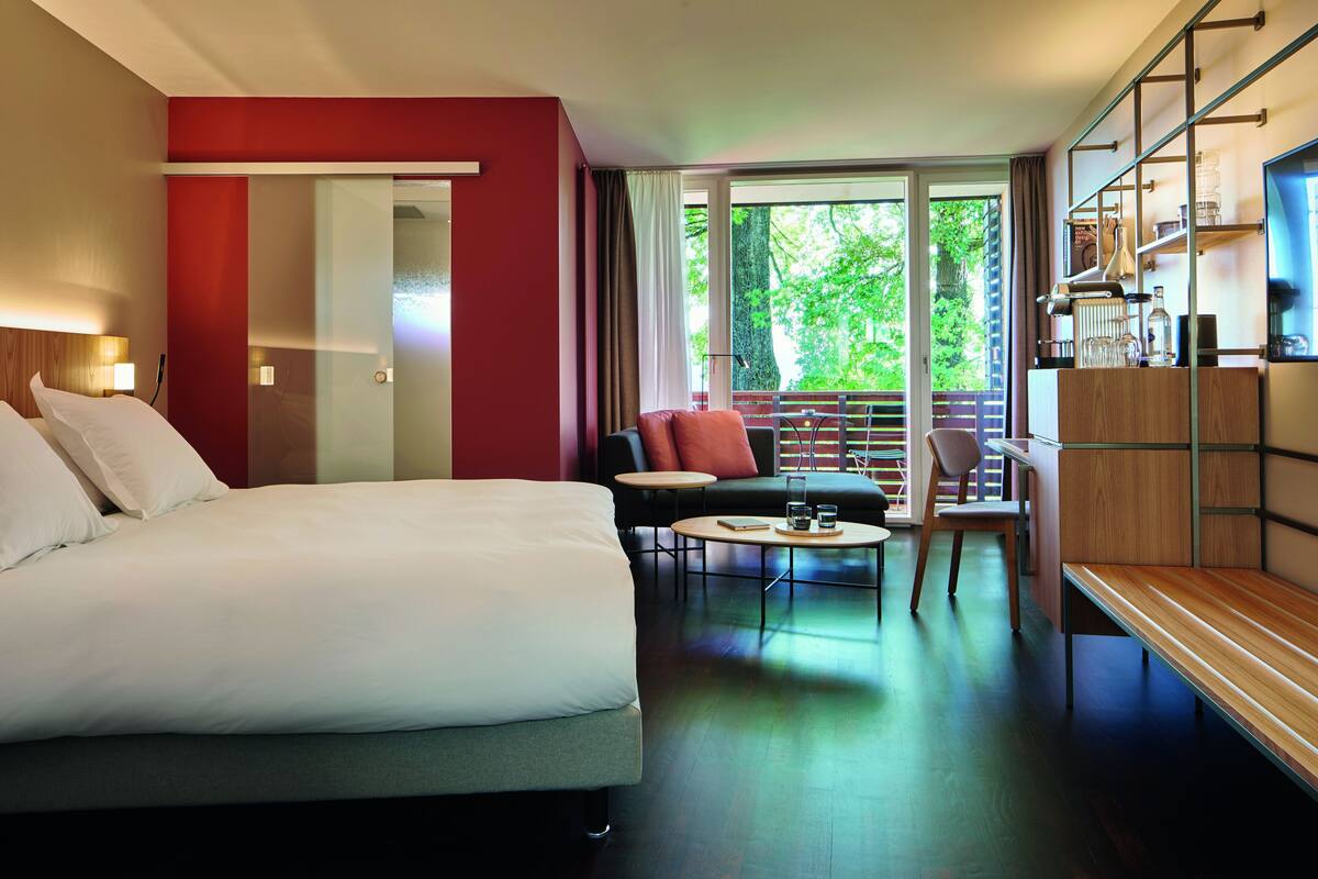Une chambre d’hôtel avec un grand lit, un canapé, une petite table et un balcon donnant sur un paysage verdoyant.