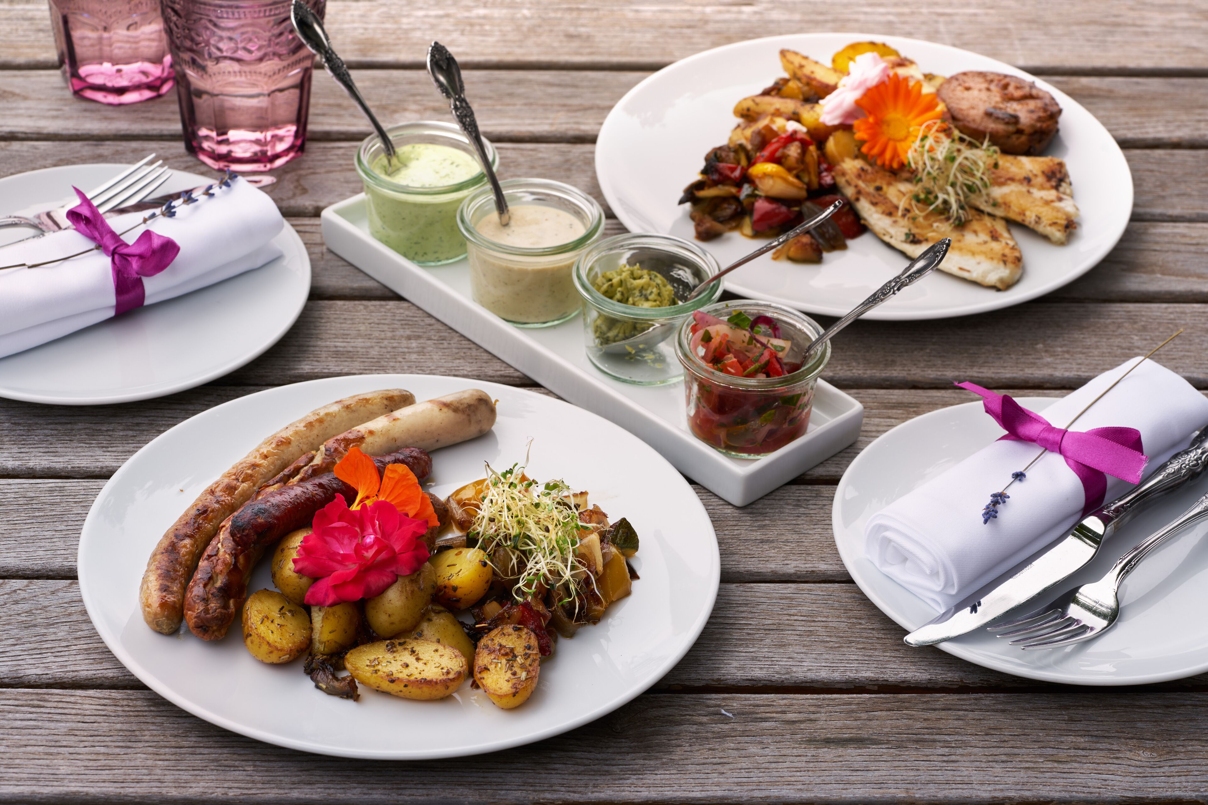 A wooden table with plates of food, including sausages, potatoes, and a salad, along with condiments and utensils.