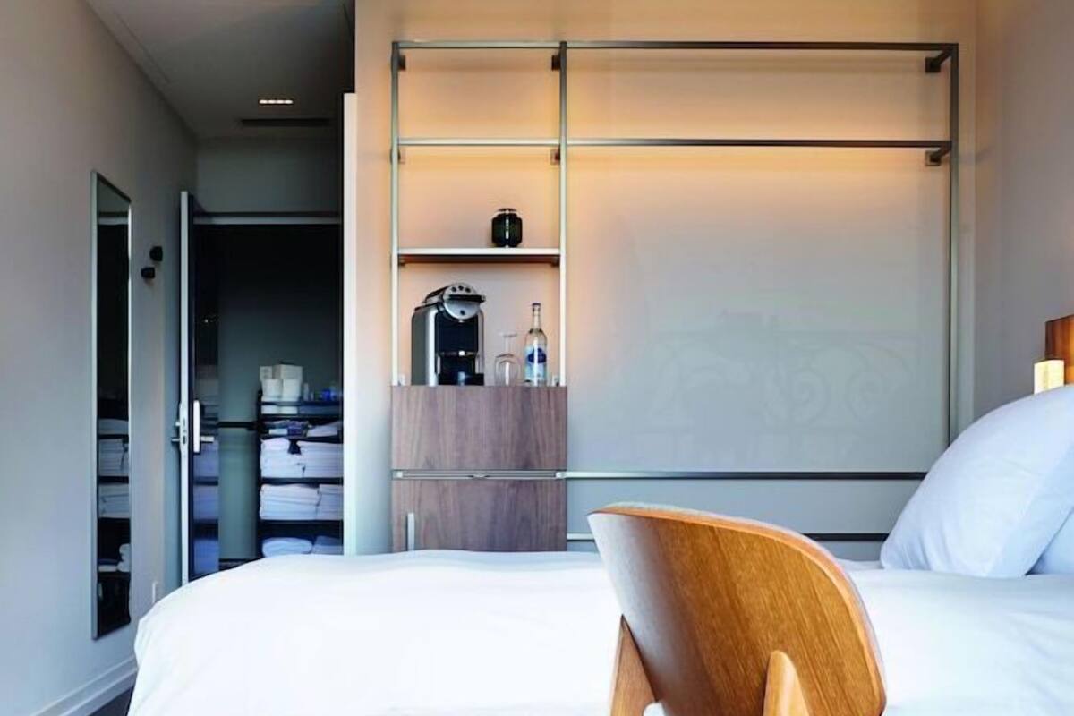 Une chambre d’hôtel moderne équipée d’un lit, d’une chaise en bois, d’une armoire et d’une petite table avec une bouilloire.