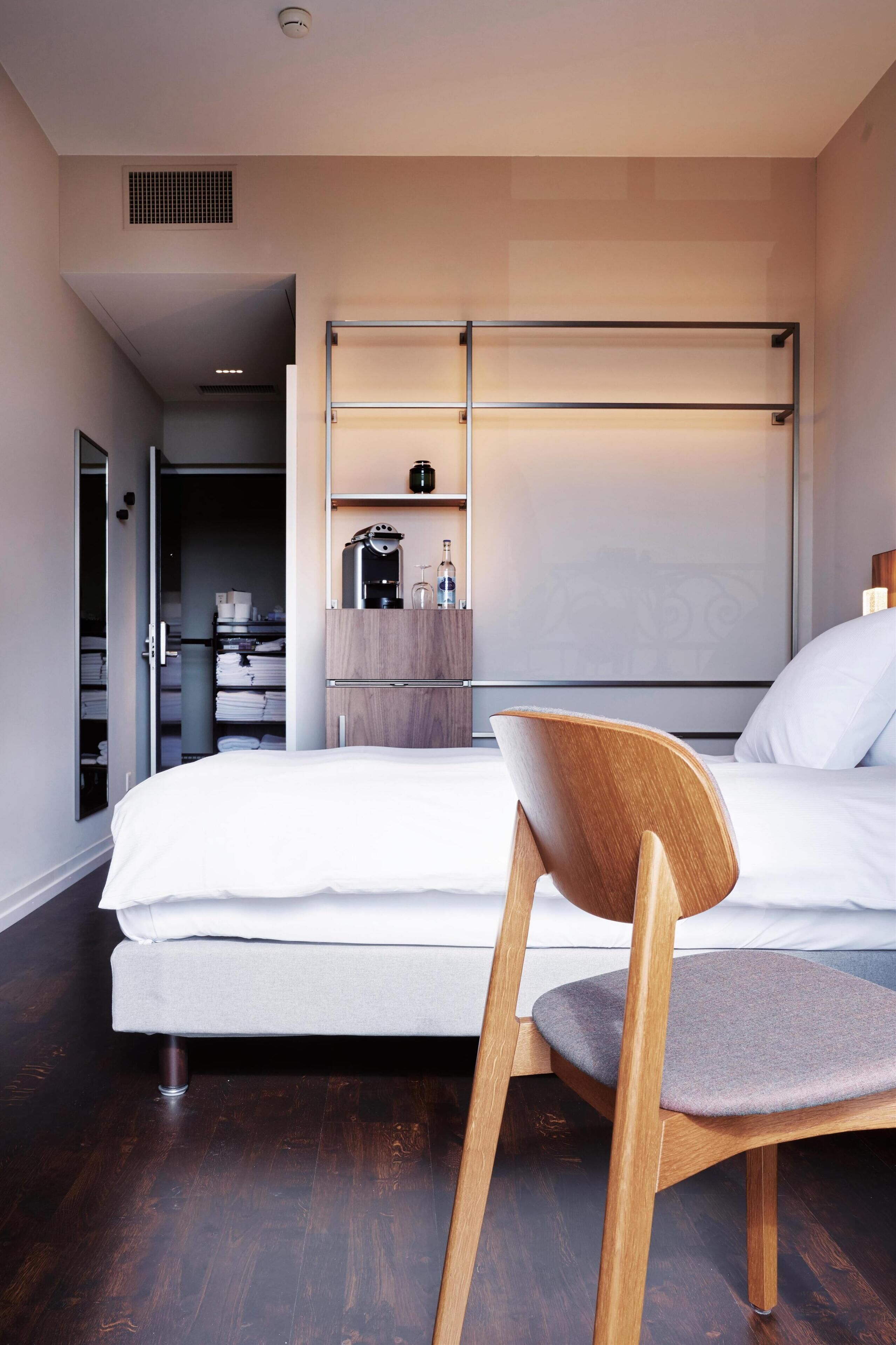 A modern hotel room with a bed, a wooden chair, a wardrobe, and a shelf with toiletries.