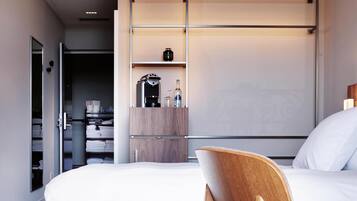 A modern hotel room with a bed, a wooden chair, a wardrobe, and a shelf with toiletries.