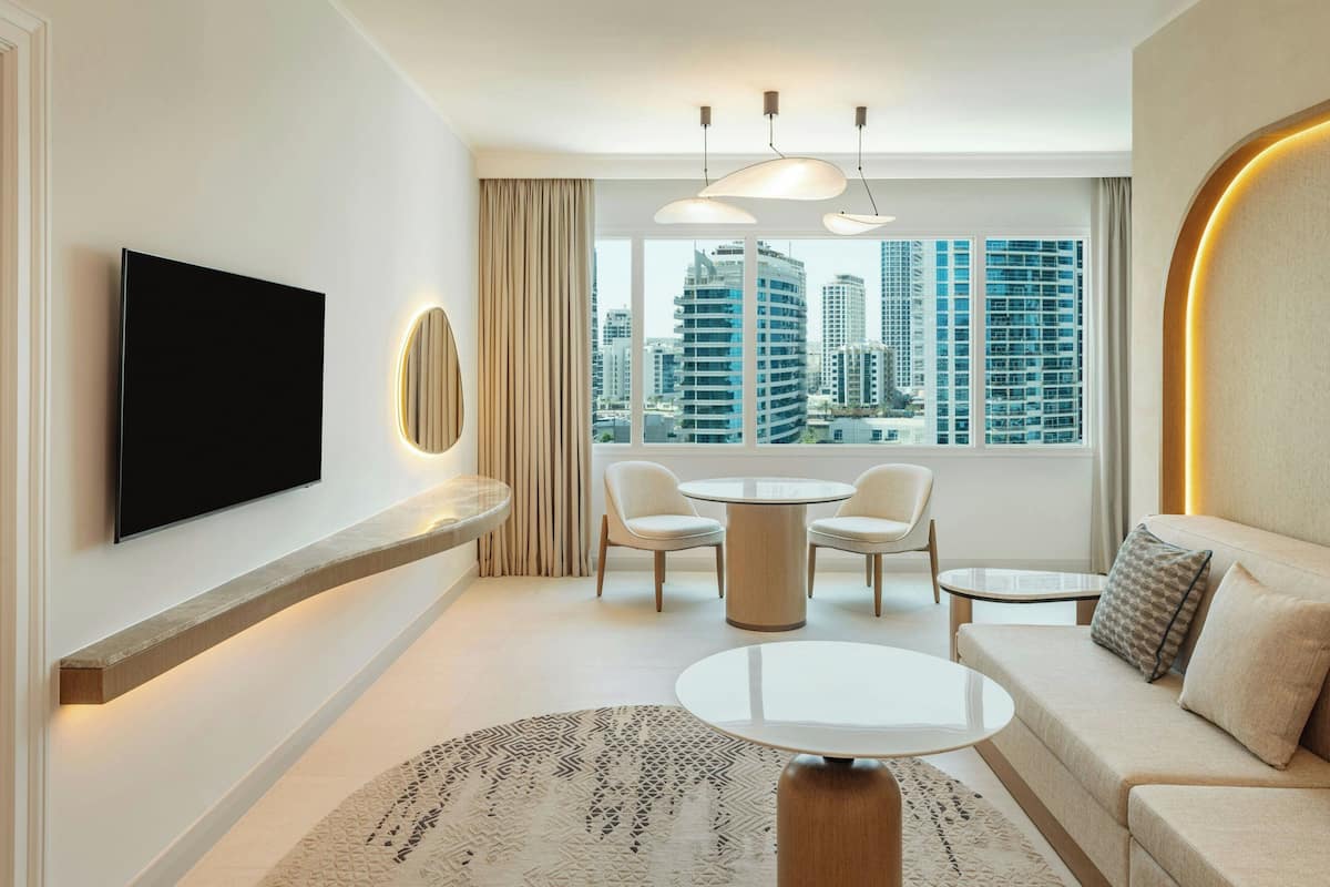A modern hotel room with a large window, a flat-screen TV, a round dining table, a sofa, and a patterned rug.