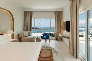 Una camera d'albergo moderna con un letto grande, un angolo salotto con una poltrona blu, una TV a parete e vista su una ruota panoramica e sulla spiaggia.