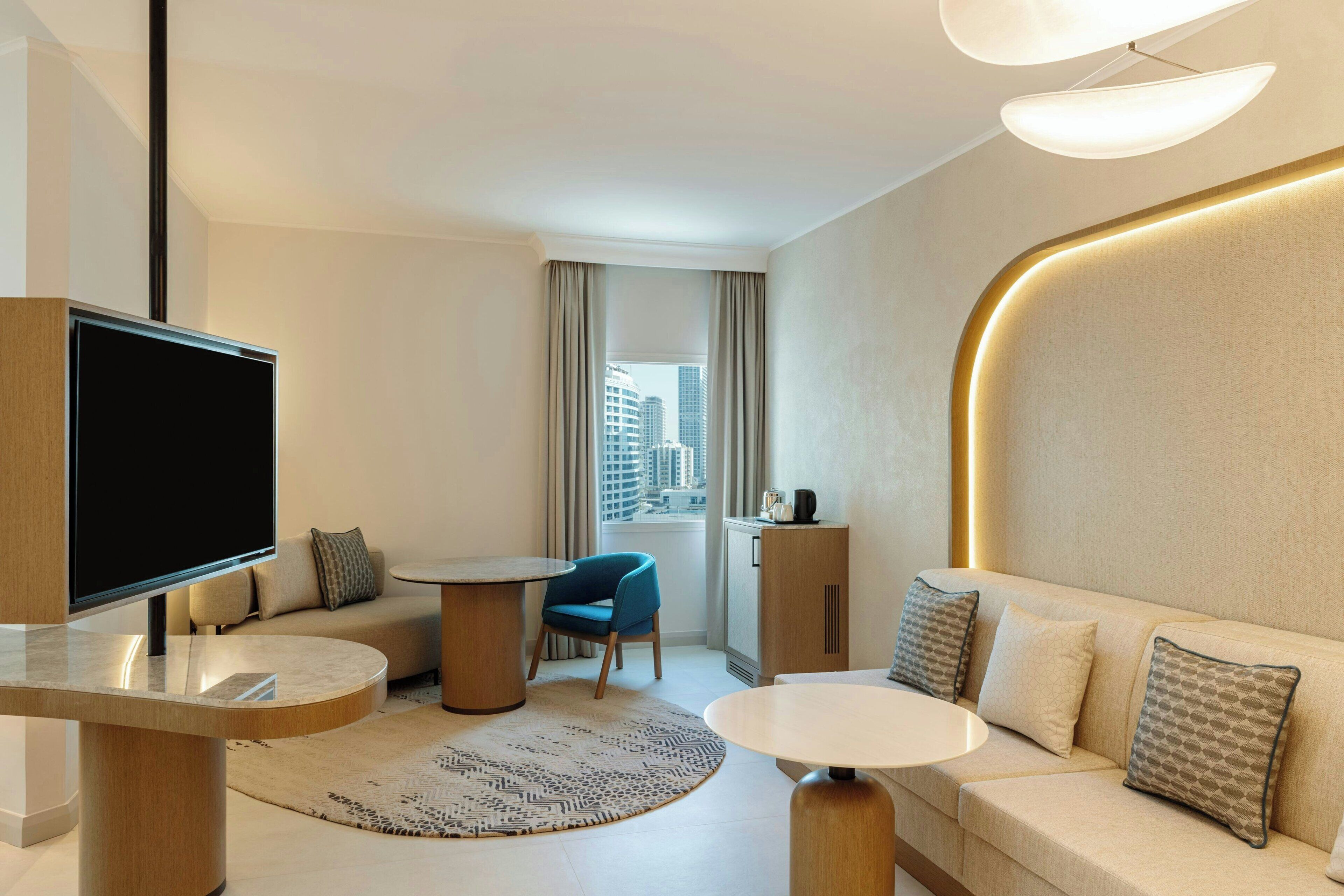 A modern hotel room with a large flat-screen TV, a sofa, a dining table, and a view of the cityscape through the window.