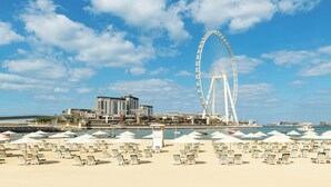 A beach with sun loungers and umbrellas, a large Ferris wheel, and a building in the background.