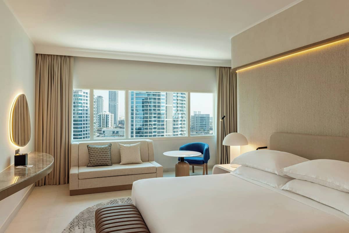 A hotel room with a bed, a sofa, a round table, and a chair. There is a window with a city view.