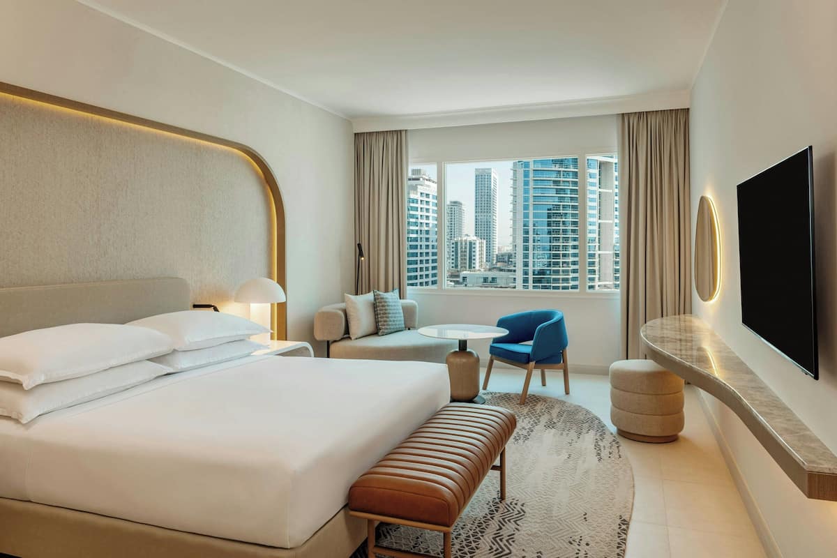 A modern hotel room with a large bed, a seating area with a table and chairs, a flat-screen TV mounted on the wall, and a view of the city skyline through large windows.