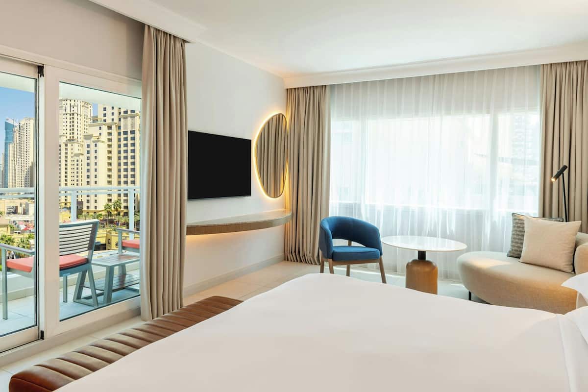 A hotel room with a large bed, a TV mounted on the wall, a balcony with a view of tall buildings, and a seating area with a table and chairs.