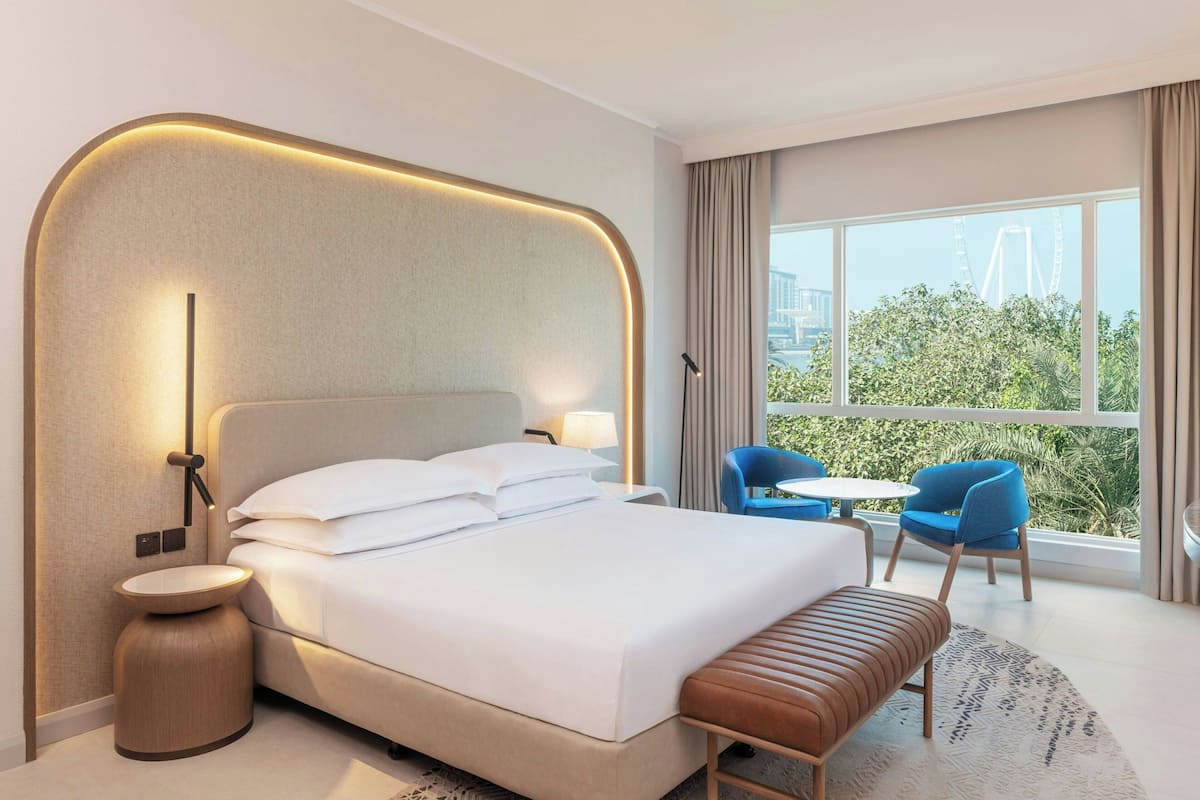A modern hotel room with a large bed, a round bedside table, a bench, a small table with chairs, and a large window with a view of greenery and a bridge.
