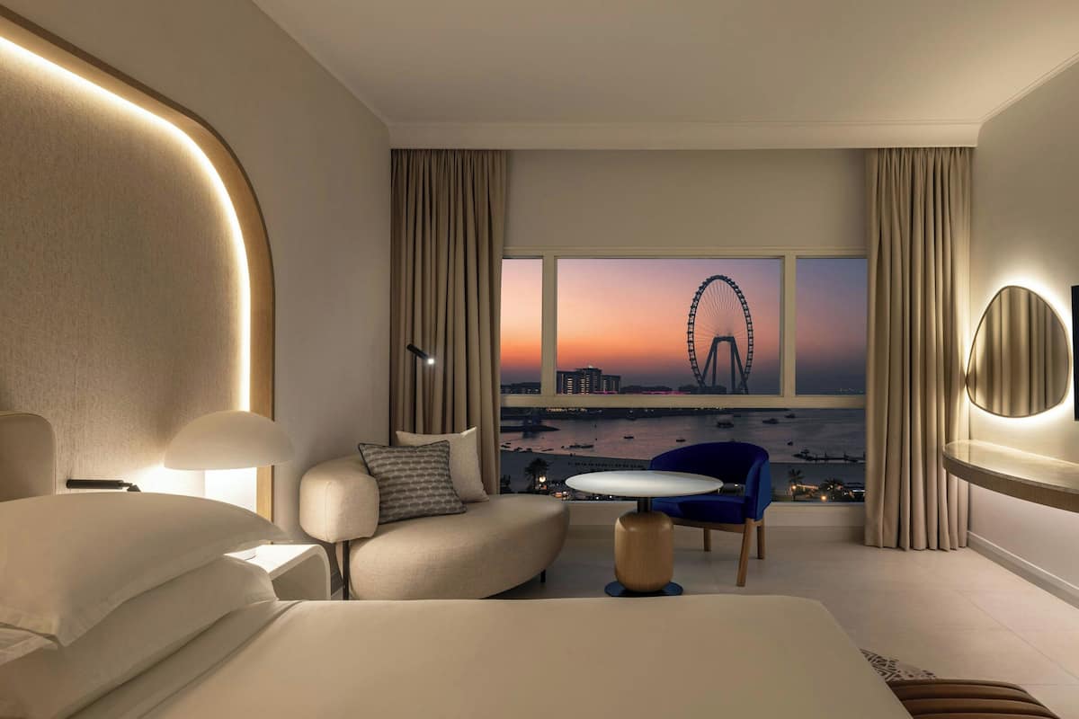 A modern hotel room with a large bed, a sofa, a small table, and a view of a Ferris wheel at sunset.