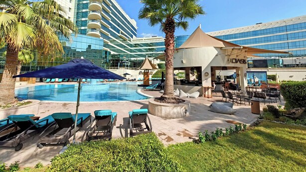 Zona de piscina de hotel con sillones de descanso, una palmera y un edificio de fachada de vidrio.
