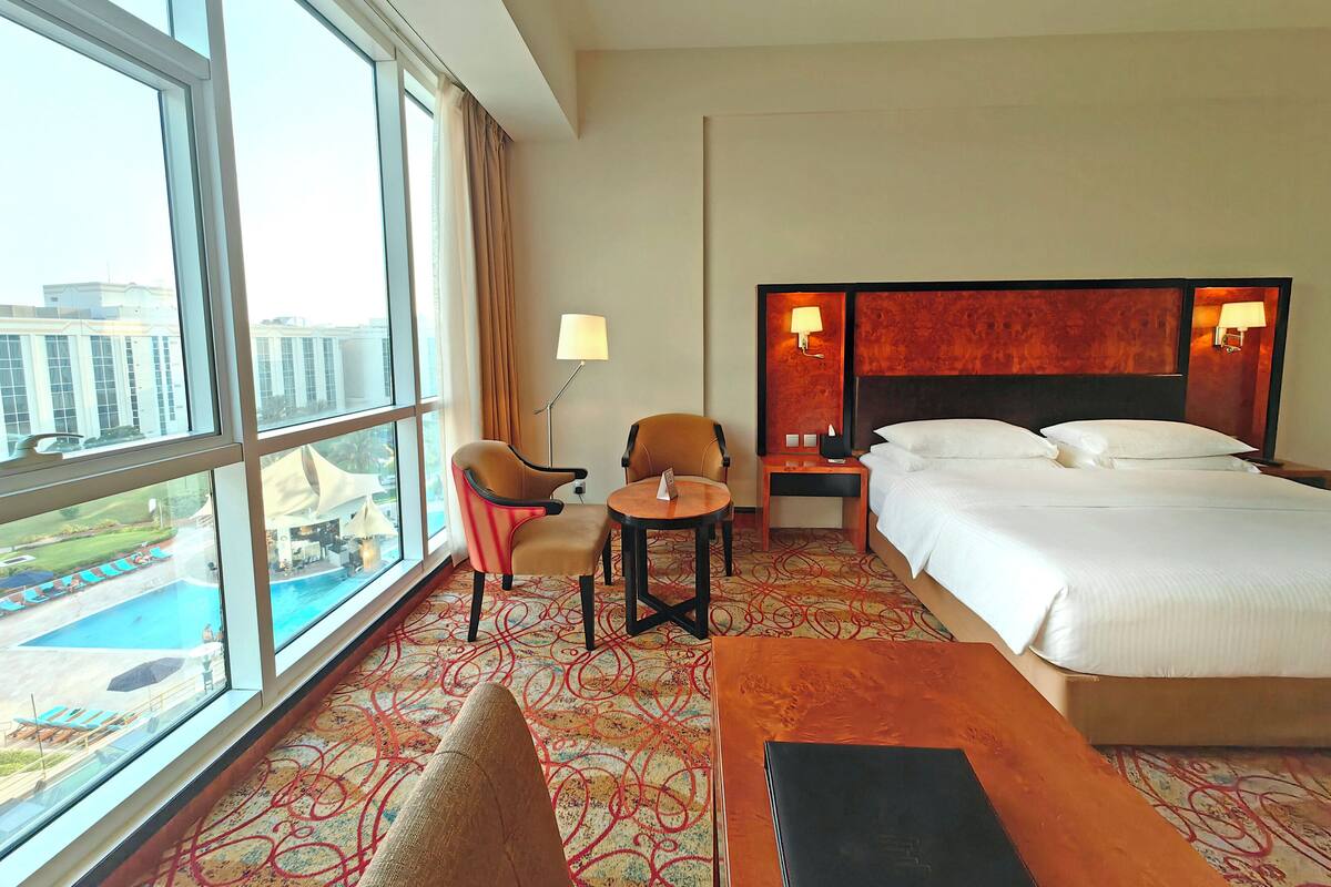 A hotel room with a large window, a bed, a desk, two chairs, a small table, and a view of a pool and buildings.