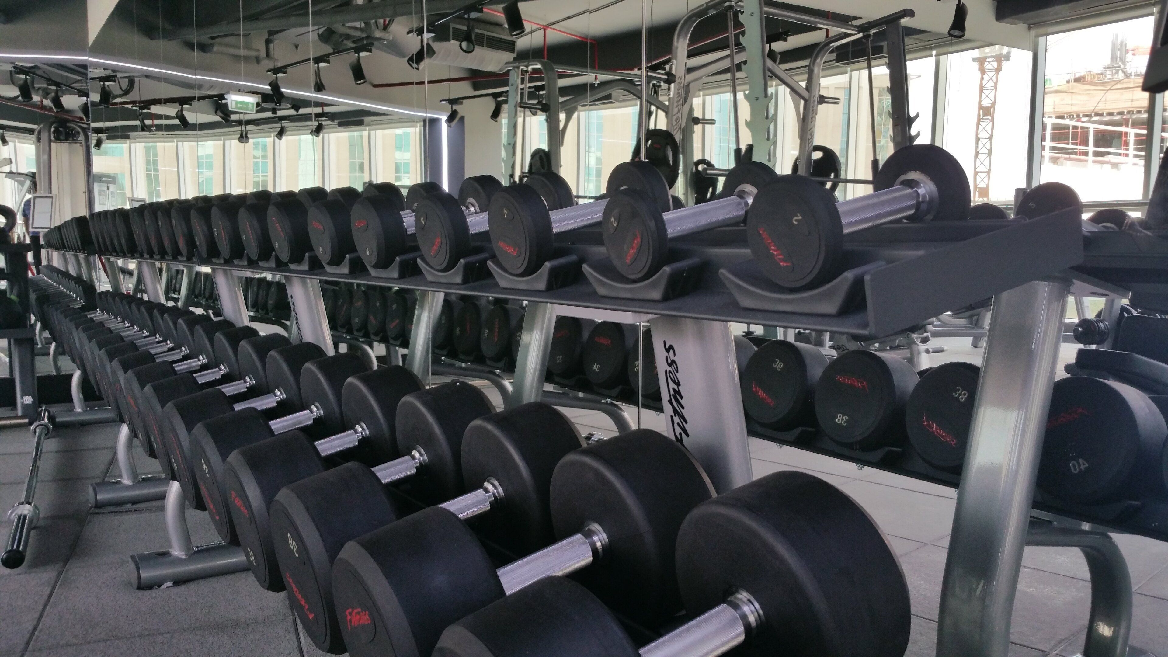 Un gimnasio con filas de mancuernas en estantes, una banca para levantamiento de pesas y un espejo grande que refleja el equipo y las ventanas.