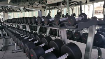 Un gimnasio con filas de mancuernas en estantes, una banca para levantamiento de pesas y un espejo grande que refleja el equipo y las ventanas.
