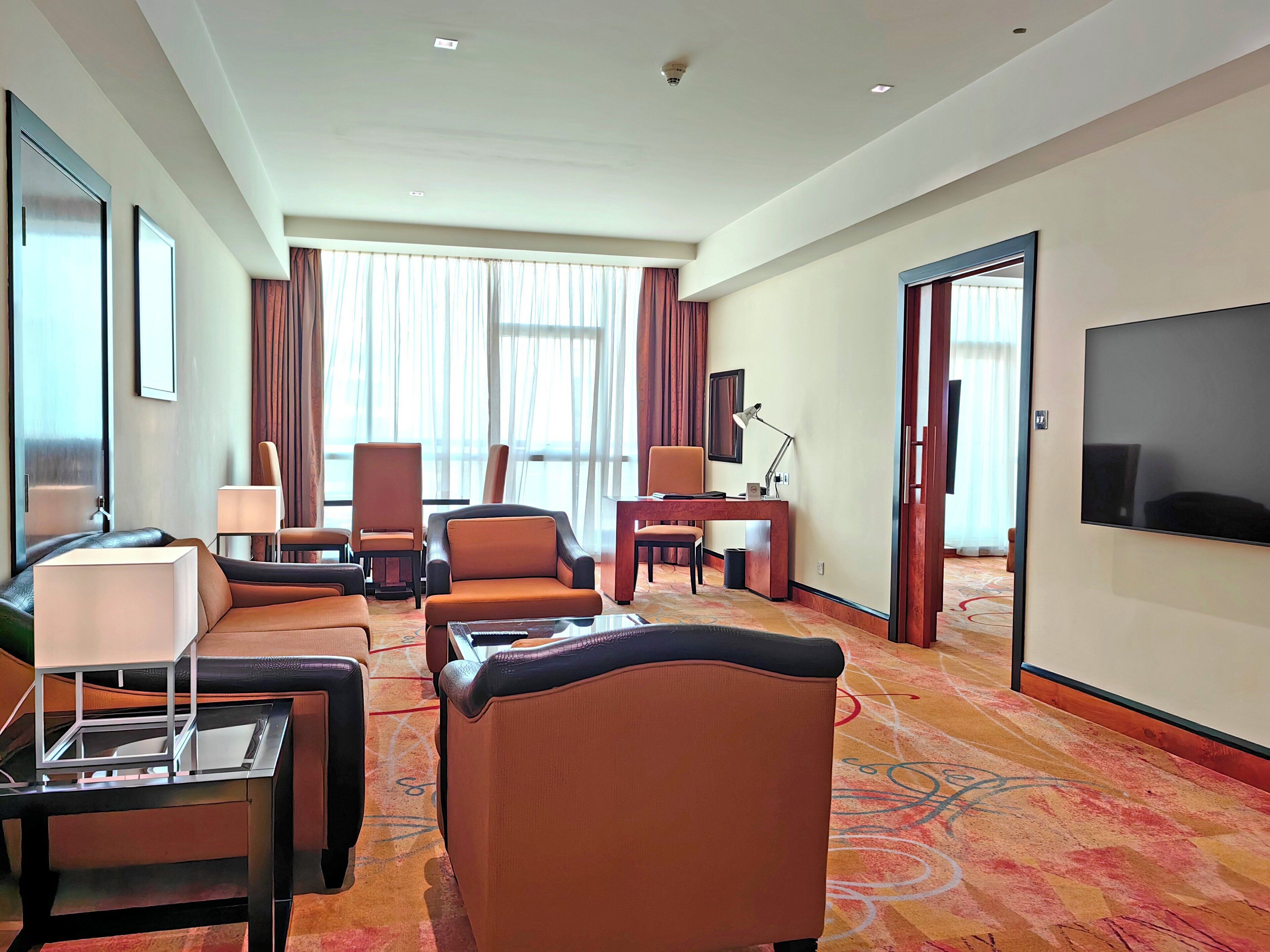 A hotel room with a sofa, armchairs, a desk with a lamp, a television, and a large window with curtains.
