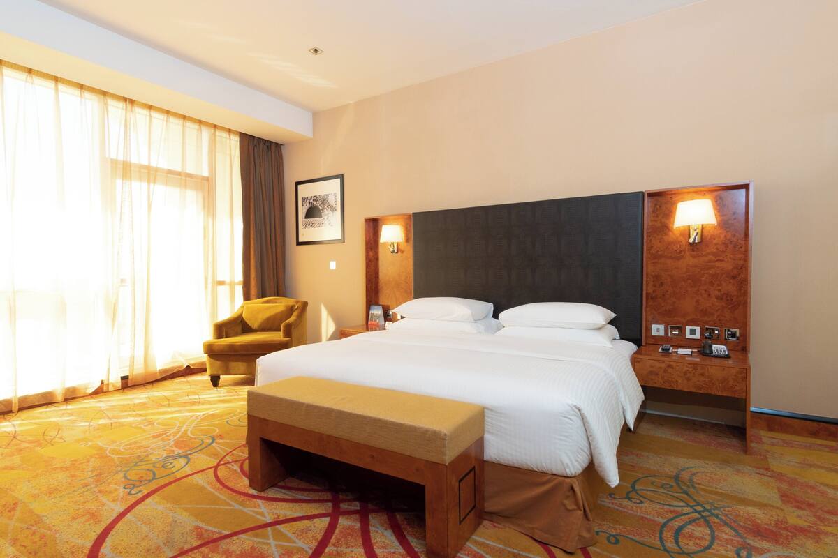 A hotel room with a large bed, a bench, a chair, and a desk with a lamp.