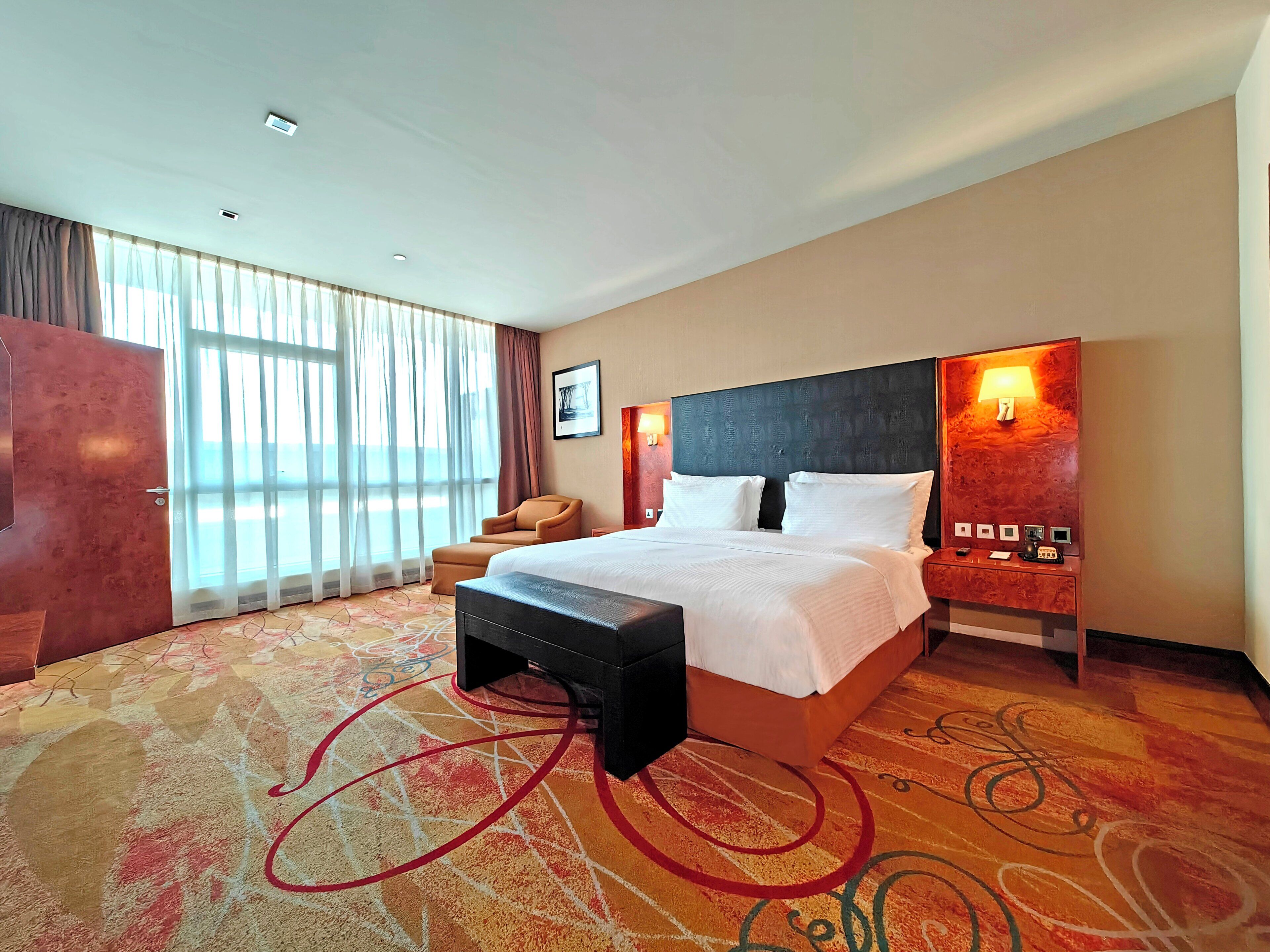 A hotel room with a large bed, a desk, and a chair. There is a window with curtains, a wall-mounted lamp, and a picture on the wall.