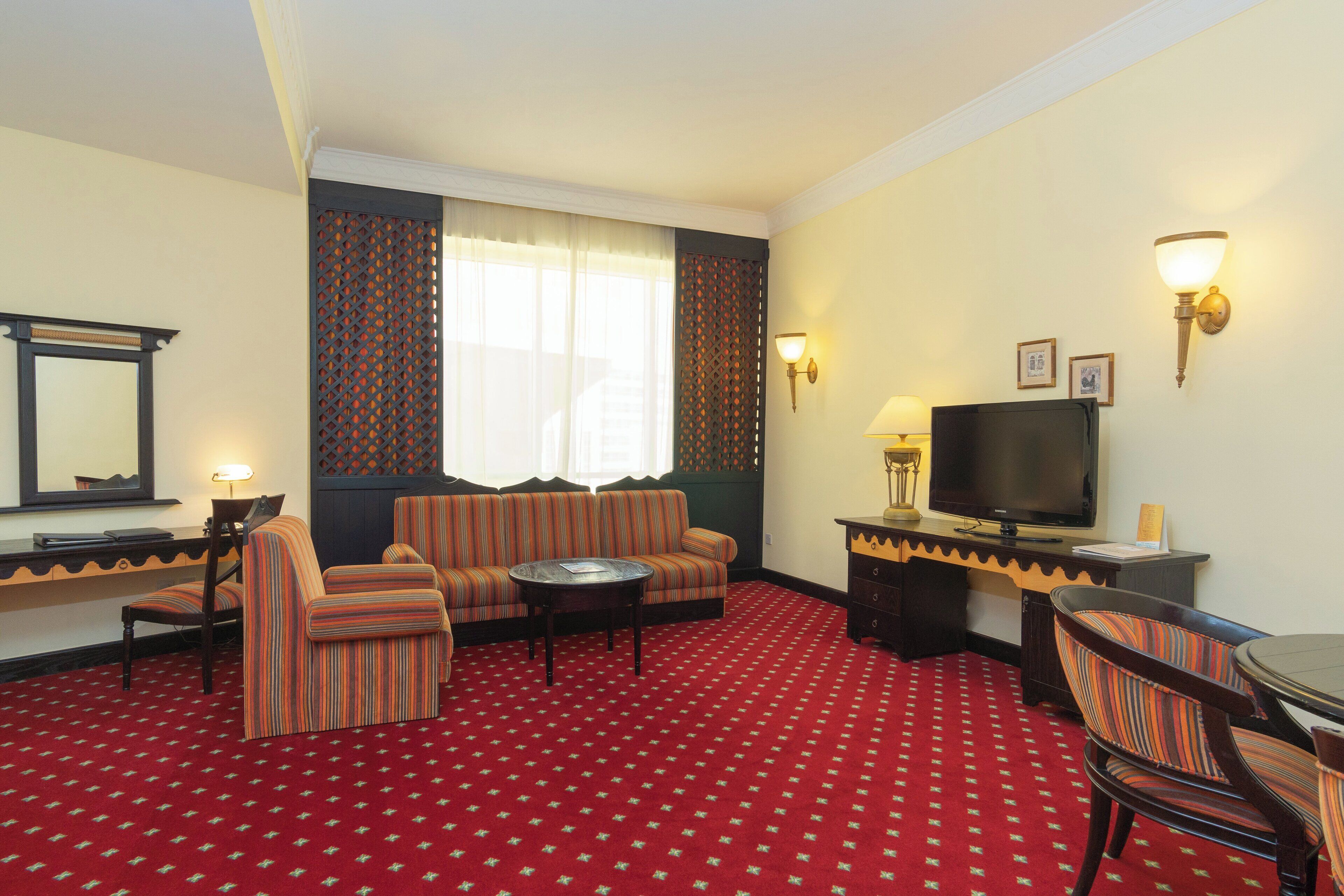 A hotel room with a red carpet, striped sofa, armchairs, a TV, and wall sconces.