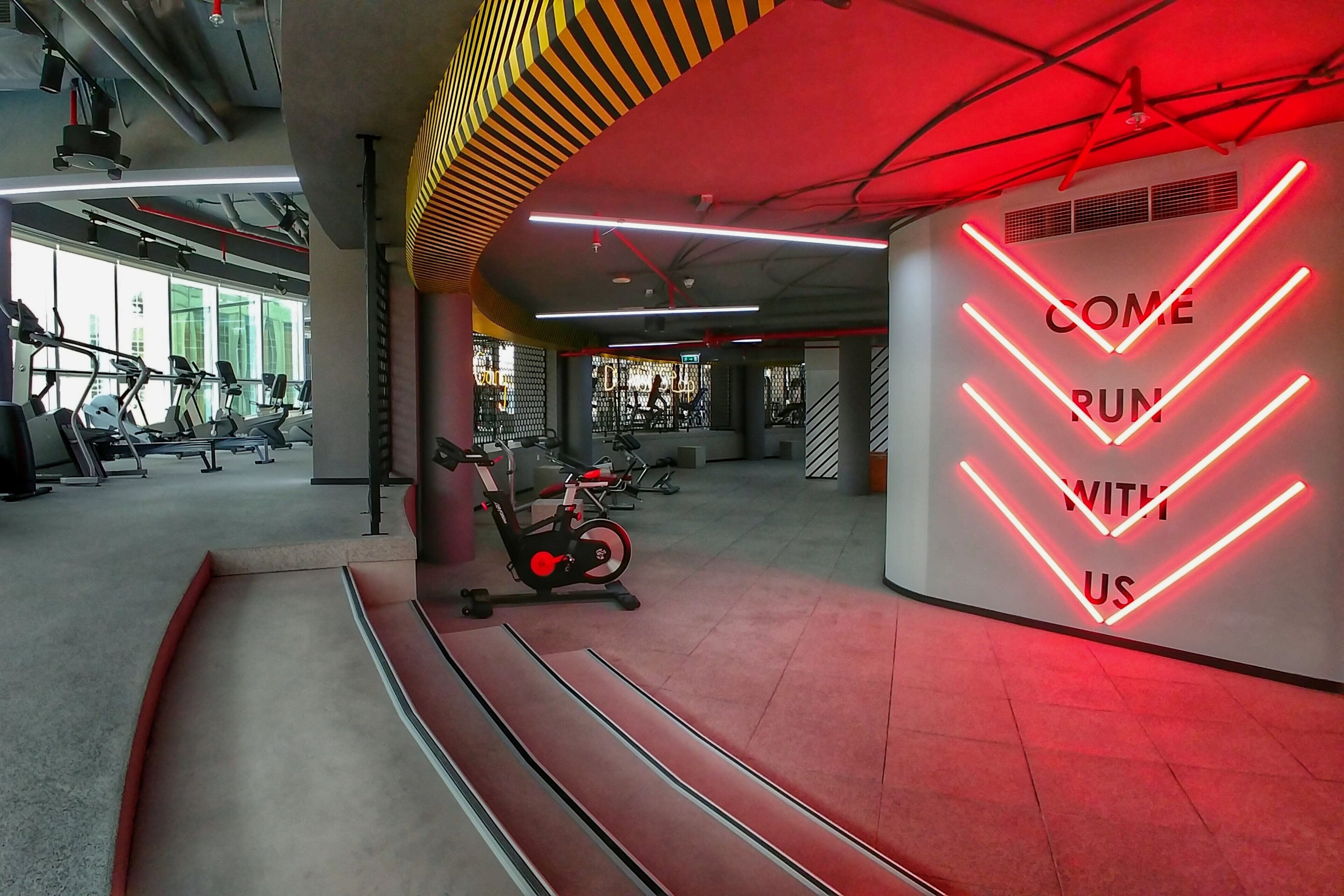 A gym with exercise equipment, a neon sign on the wall, and a red and yellow ceiling design.
