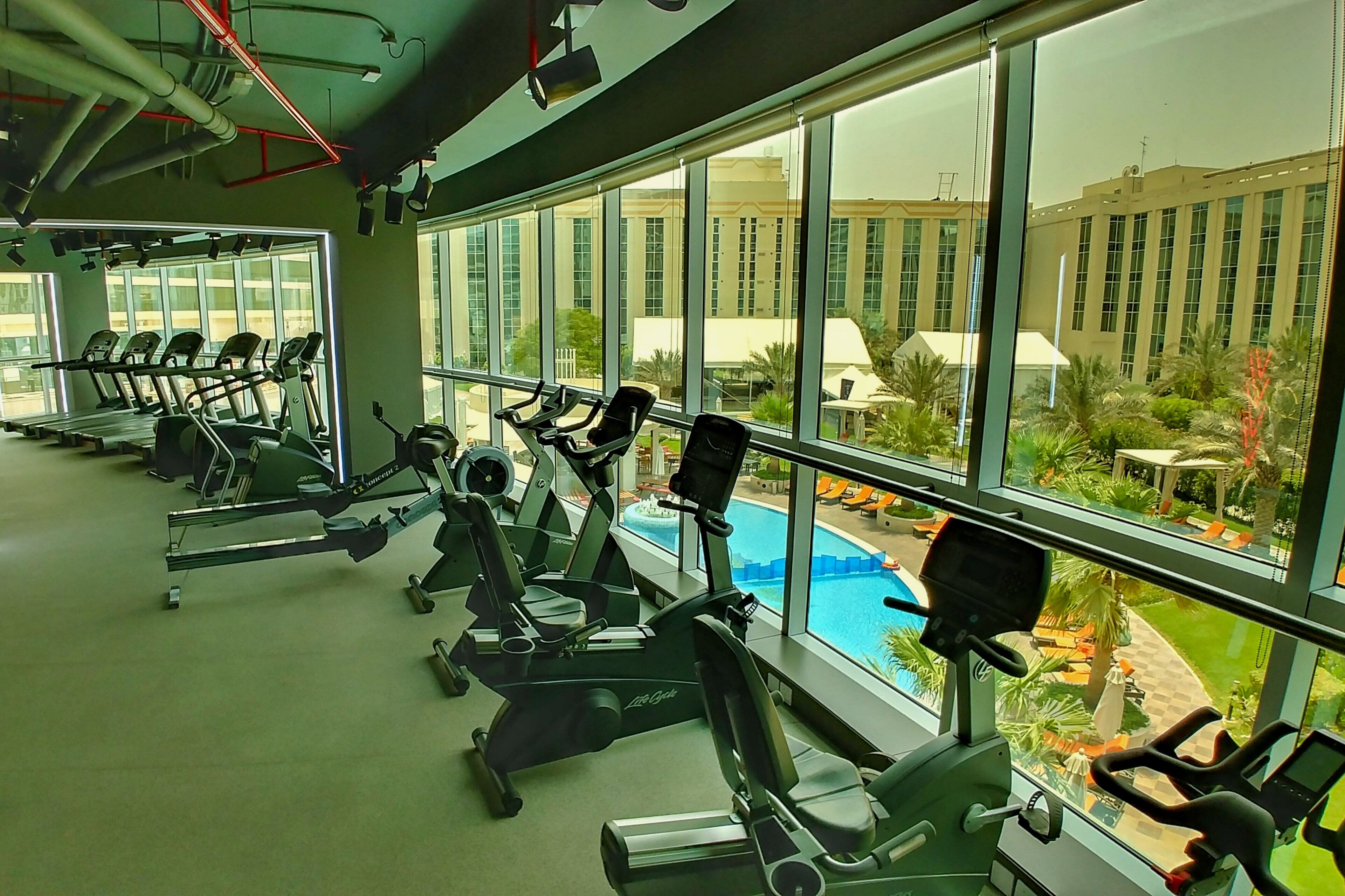 Un gimnasio con equipo de ejercicio y amplias ventanas con vistas a una piscina y un edificio.