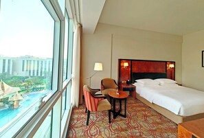 Ein Hotelzimmer mit einem Bett, zwei Stühlen, einem kleinen Tisch, einer Lampe und Blick auf ein Gebäude und Palmen durch große Fenster.