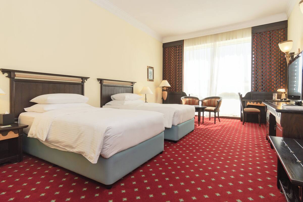 A hotel room with two beds, a desk, a chair, and a TV.
