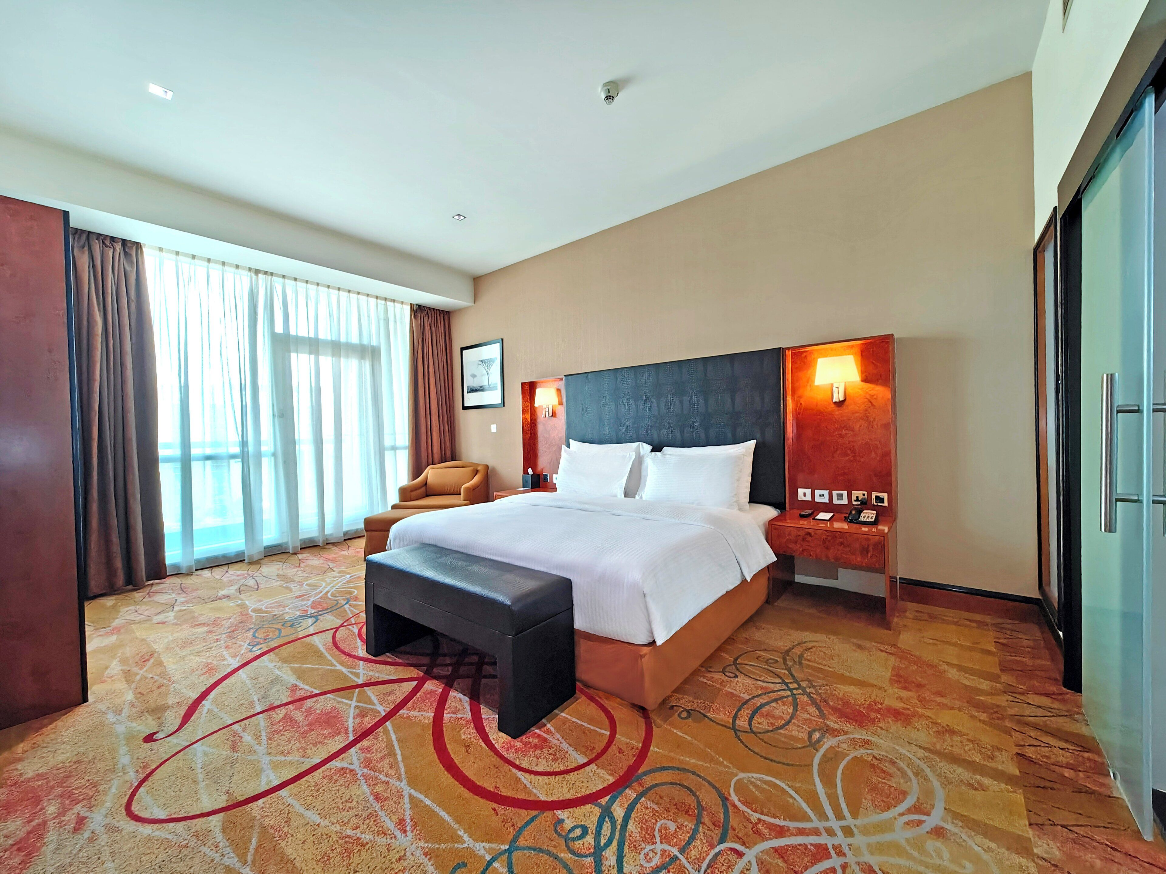 A hotel room with a large bed, a desk with a lamp, a chair, and a large window with curtains.