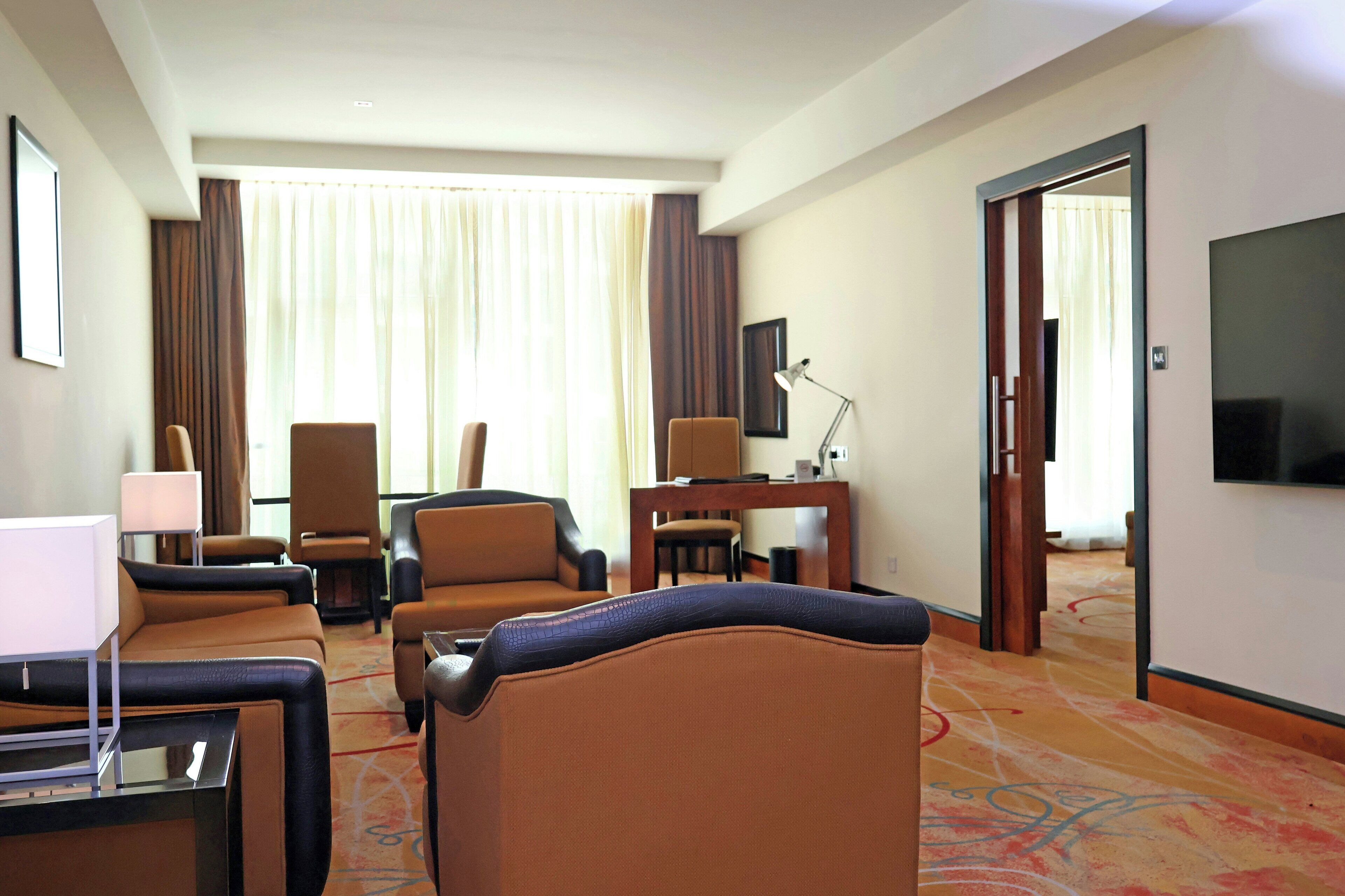 Quarto de hotel com área de estar, mesa de trabalho, televisão e uma janela grande com cortinas.