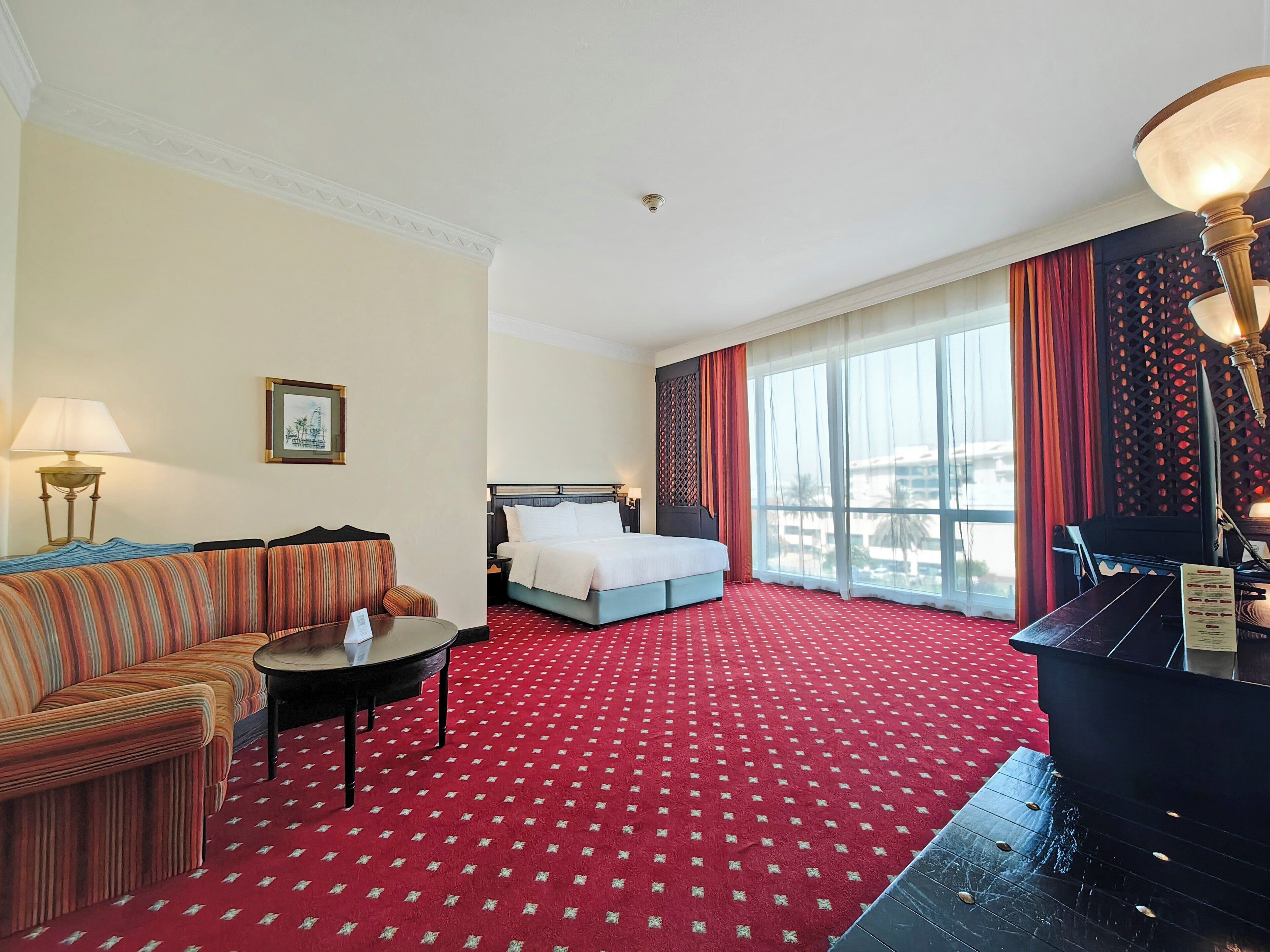 A hotel room with a red carpet, a bed, a sofa, a small table, and a piano.