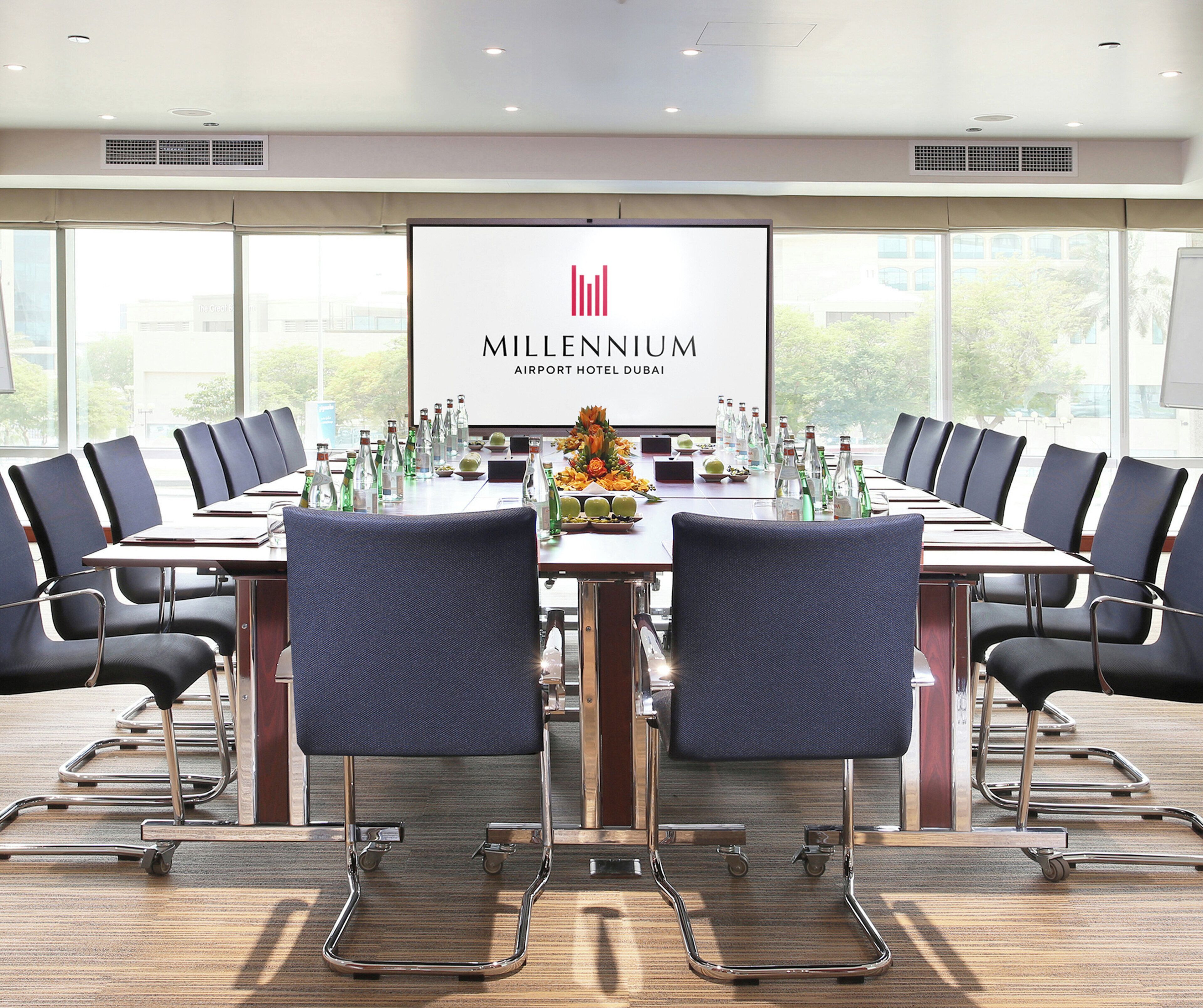Une salle de conférence à l’hôtel Millennium Airport de Dubaï, avec une longue table, des chaises et un grand écran affichant le nom de l’hôtel.