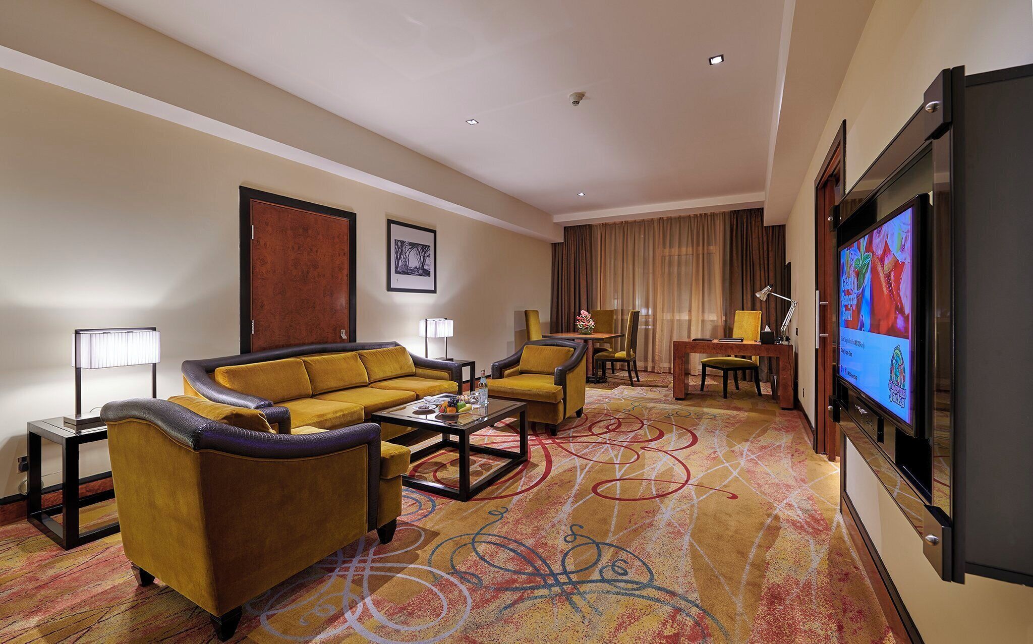 Una habitación de hotel con sofá, sillones, una mesa de centro, televisor y zona de comedor con mesa y sillas.