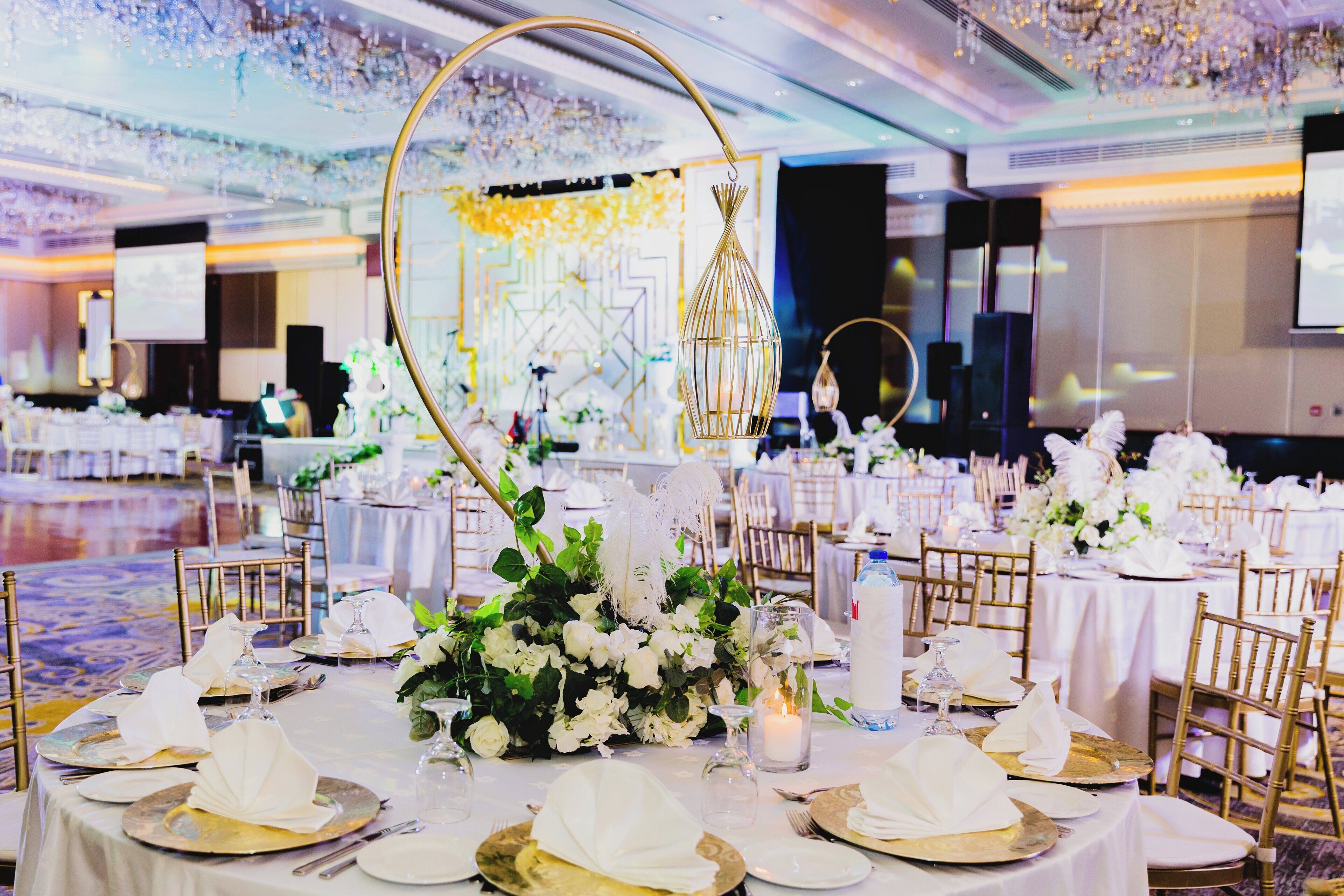 A banquet hall with round tables set for a formal event, featuring gold chairs, white tablecloths, and floral centerpieces.