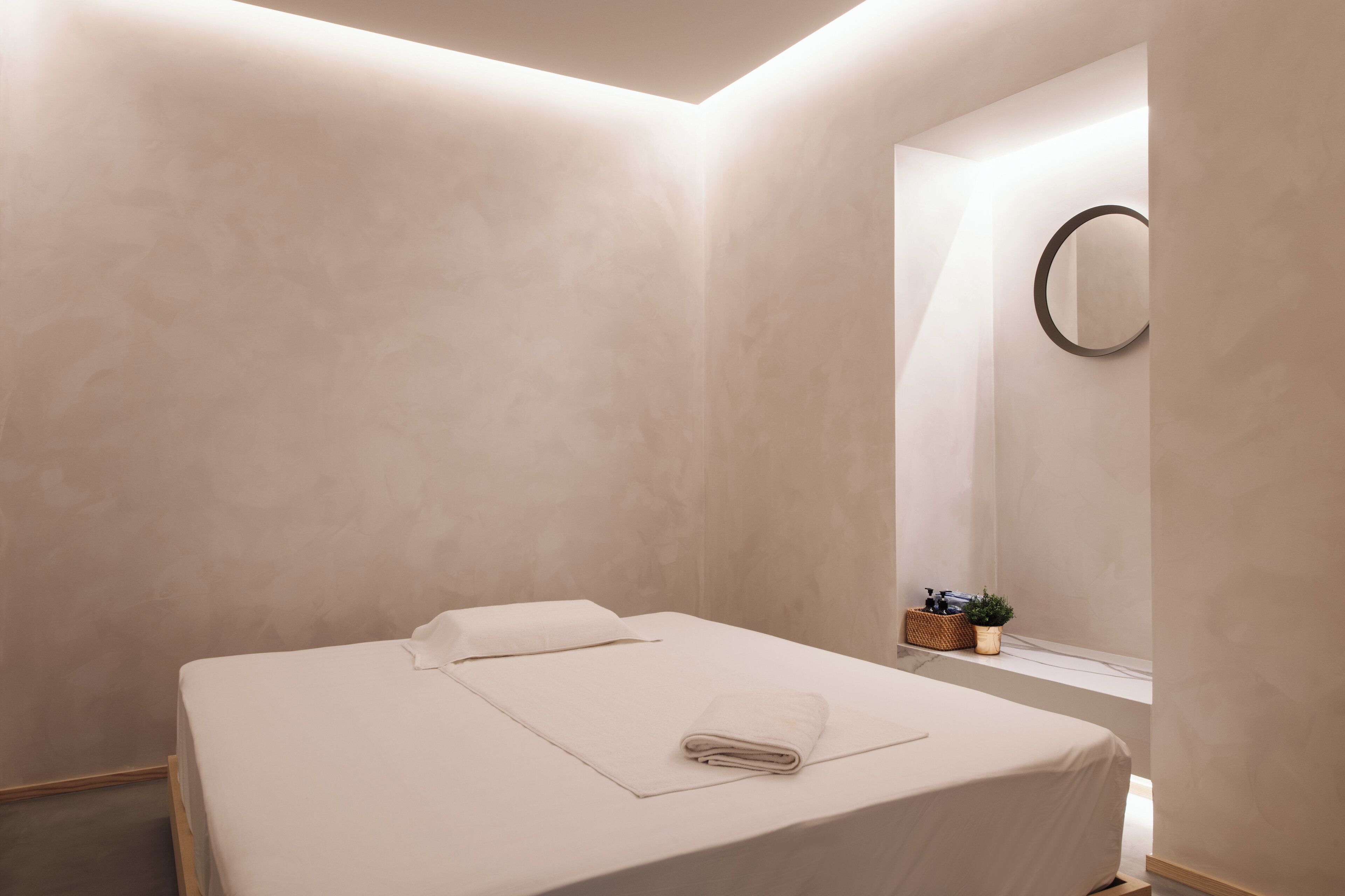 A minimalist hotel room with a bed, a small window, and a wall-mounted mirror.