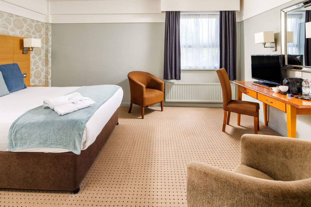 Ein Hotelzimmer mit einem Bett, zwei Sesseln, einem Schreibtisch mit einem Computer und einem großen Spiegel.