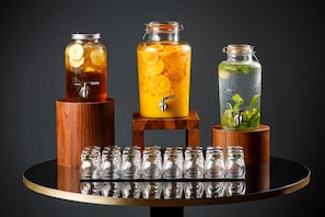 Trois bocaux en verre contenant différentes boissons, placés sur un présentoir en bois, et une rangée de bouteilles en verre vides en dessous.