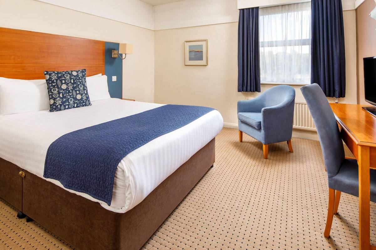 Ein Hotelzimmer mit einem großen Bett, einem Schreibtisch, einem Stuhl, einem Fernseher und einem Vorhangfenster.