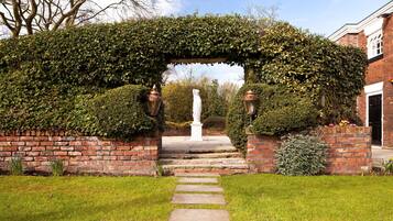 A brick pathway leading to a white statue under a vine-covered archway.