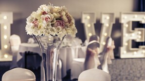Une table dressée pour un événement formel, agrémentée d’un grand vase rempli de fleurs, d’un miroir illuminé et d’un panneau portant le mot « AMOUR » en arrière-plan.
