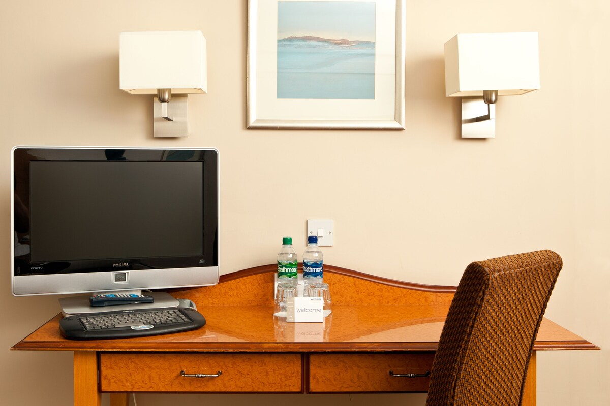 Een hotelkamer met een bureau, een flatscreen-tv, een toetsenbord, een afstandsbediening, twee waterflesjes, een ingelijste foto aan de muur en twee wandlampen.