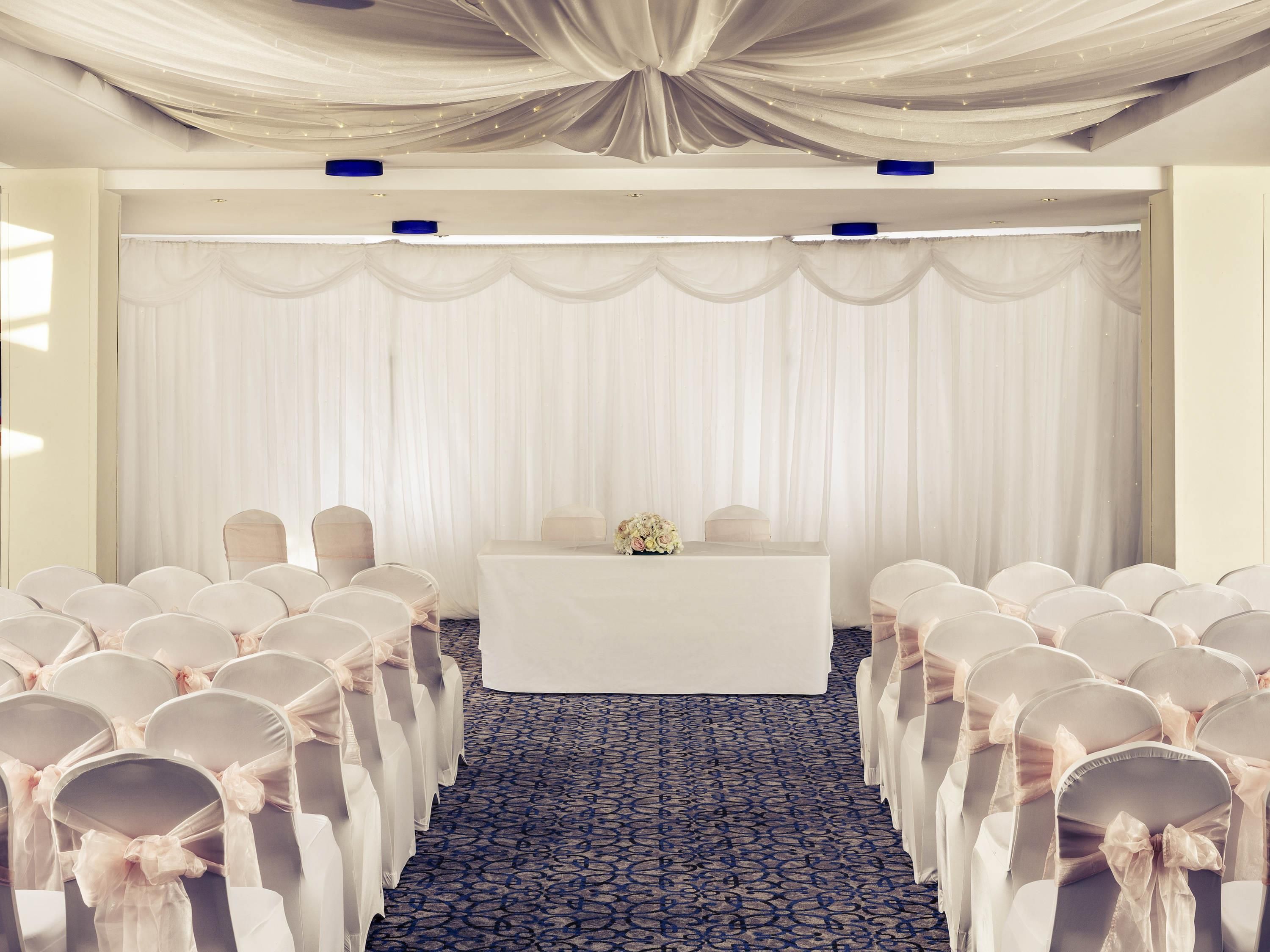 Un salón de conferencias con filas de sillas, una mesa con un arreglo floral y cortinas decorativas.