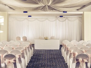 Un salón de conferencias con filas de sillas, una mesa con un arreglo floral y cortinas decorativas.