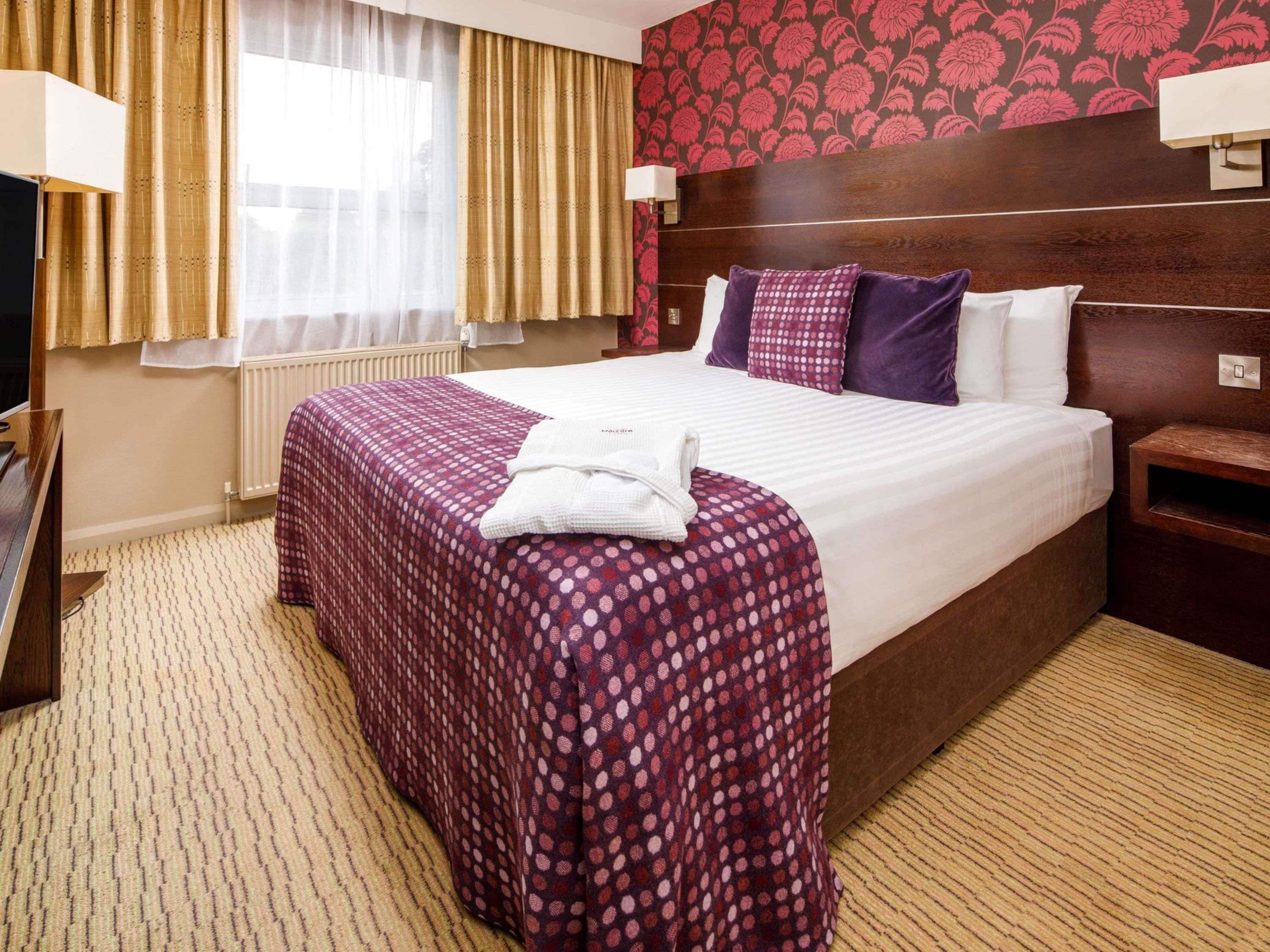 A hotel room with a large bed, a wooden headboard, a window with curtains, a radiator, and a bedside table with a lamp.